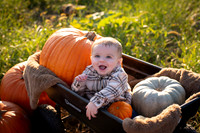Smiling baby sitting in a wagon surrounded by large pumpkins during a fall pumpkin patch family photography session in Virginia.